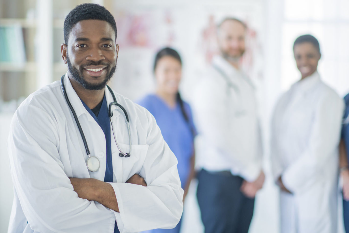 A group of doctors and nurses are standing together in a medical office. One stands in front of the others as they all look forward and smile.