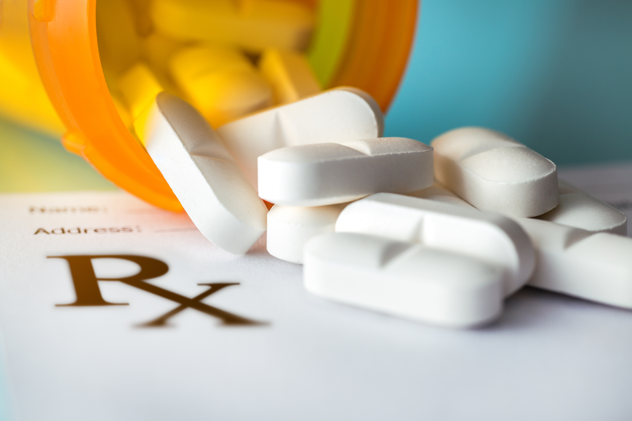 Prescription drug bottle on its side with white pills spilling out over a piece of paper with Rx symbol on it.